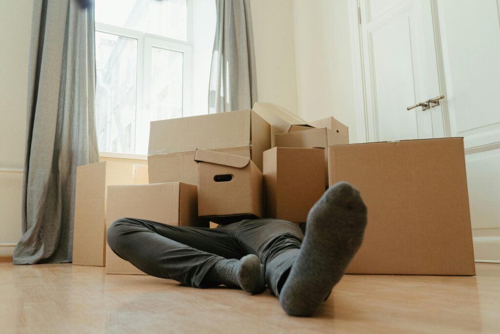 A person lies down tired among cardboard boxes in a bright room, depicting moving day stress.