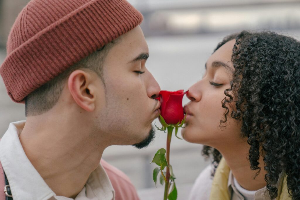 Side view of romantic Hispanic couple with closed eyes kissing red rose while spending time on embankment against blurred background