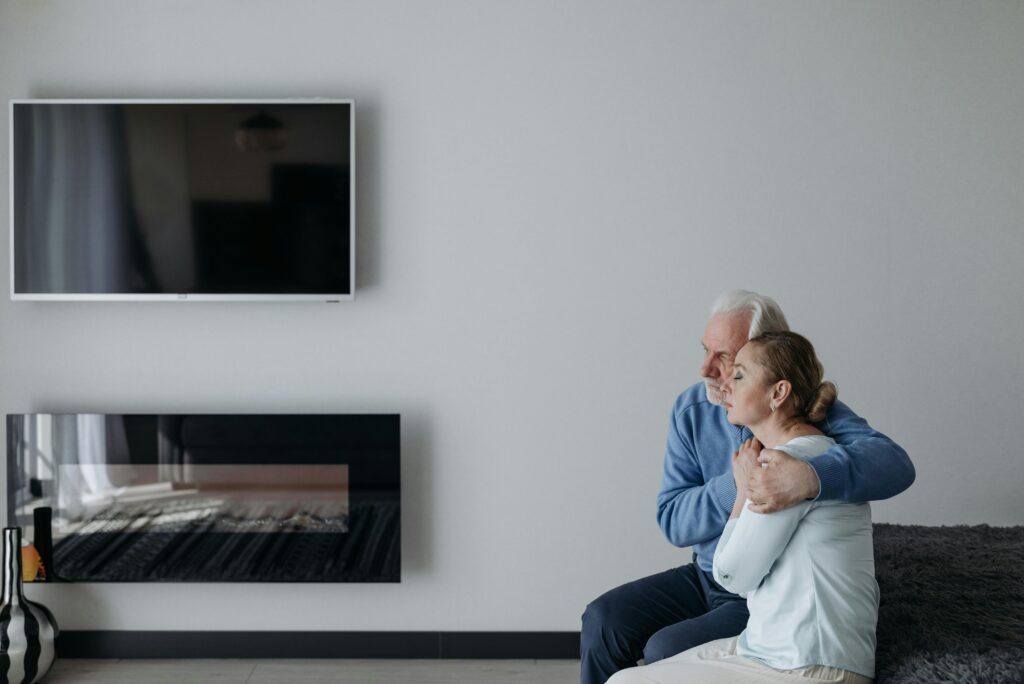An elderly couple sits on the bed, embracing and comforting each other in a modern bedroom setting.