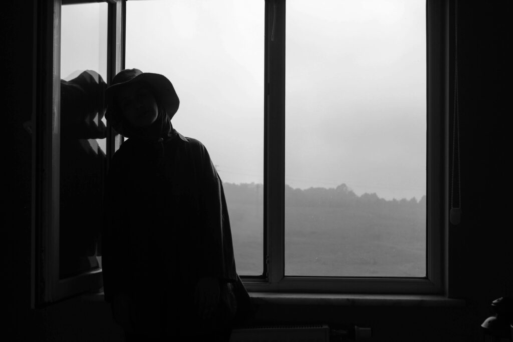 Black and white silhouette of a person wearing a hat while leaning by a window.
